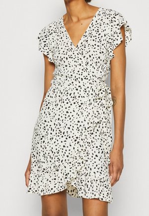 Witte wikkeljurk met zwarte onregelmatige stippen, korte vleugelvormige mouwen, geruffleerde zoom en een strikdetail in de taille, gedragen door een staande persoon.