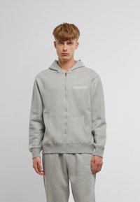 Sivi zip-up dukserica napravljen od mekog materijala, s prednjim džepom, rebrastim manžetama i logotipom na lijevoj strani prsnog područja.