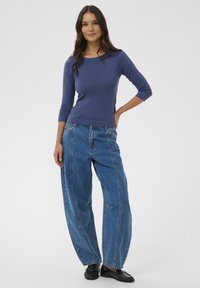 Femme debout, une main sur la hanche, portant un haut bleu à manches trois-quarts, un jean bleu à jambes larges avec des coutures verticales, et des mocassins noirs.