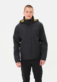 Veste imperméable noire avec une capuche bordée de jaune, fermeture éclair à l'avant et poignets ajustables, portée sur un pantalon noir. Logo Vaude sur la poitrine.