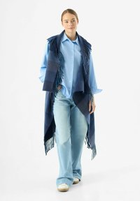 Poncho a scacchi blu e navy con orli frangiati, indossato sopra una camicia azzurro chiaro e jeans azzurro chiaro, abbinato a sneakers bianche.