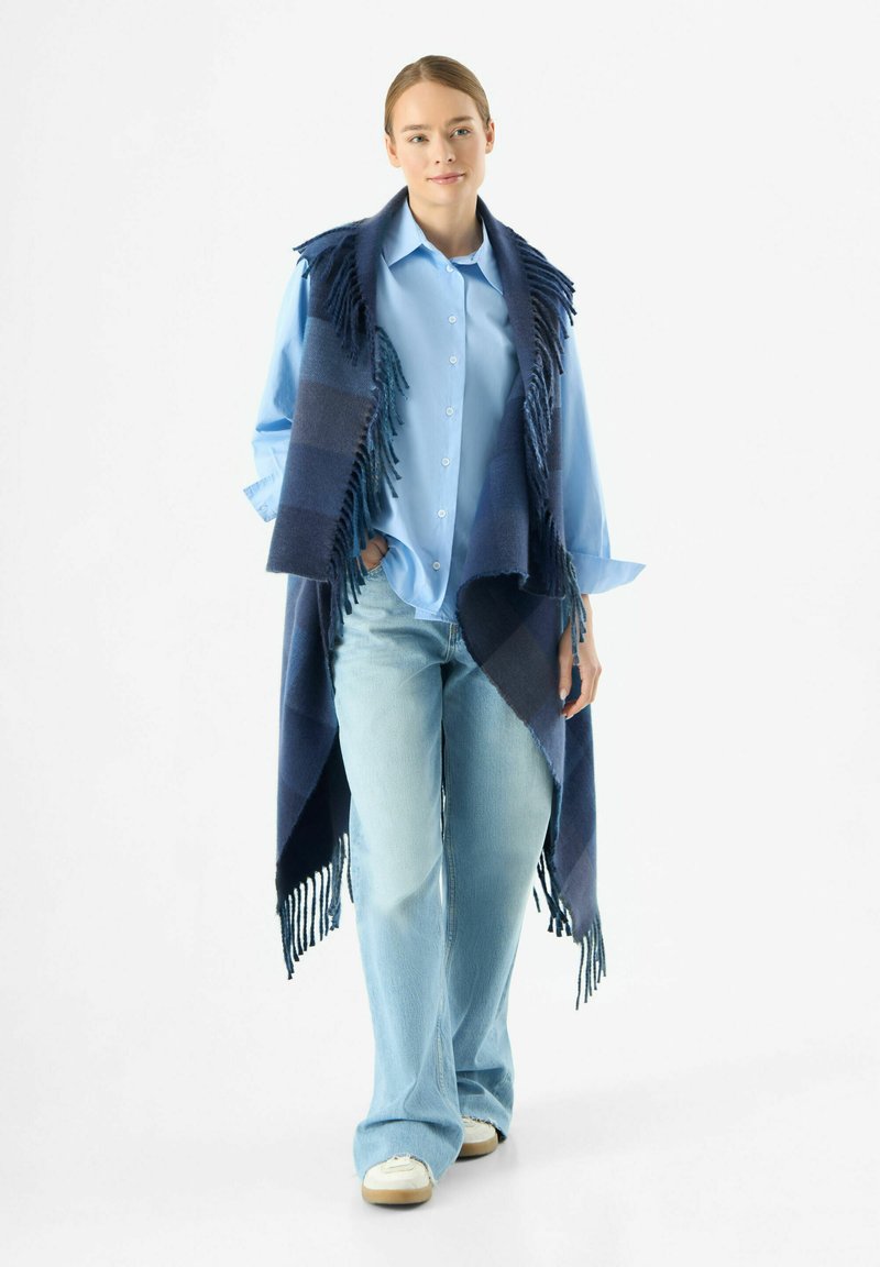 Poncho a scacchi blu e navy con orli frangiati, indossato sopra una camicia azzurro chiaro e jeans azzurro chiaro, abbinato a sneakers bianche.