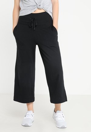 Femme portant un pantalon large noir raccourci à taille coulissante, un t-shirt gris noué et des baskets blanches, debout devant un fond blanc.
