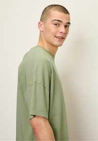 Olīvu zaļš, pārāk liels T-krekls, izgatavots no mīkstas kokvilnas, ar apaļu apkakli un īsām piedurknēm, ar vieglu tekstūru un brīvu piegriezumu.