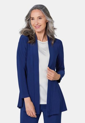 Marineblauwe cardigan met open voorkant, lange mouwen, lichtgewicht stof en een relaxed fit, gedragen over een witte top met subtiele versieringen.