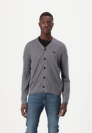 Gilet gris en tricot, avec un col en V, devant boutonné et poignet côtelé. Petite étiquette à logo sur la poitrine gauche. Jean en denim.