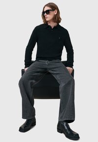 Mustapullopaita, jossa on kaulus, yhdistettynä löysiin harmaisiin farkkulegginseihin ja mustiin nilkkabootseihin. Mallilla on tummat aurinkolasit.