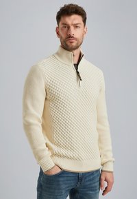 Creme-kleurige gebreide sweater met een ritssluiting op de kraag, voorzien van een gestructureerd ruitpatroon en ribbels aan de manchetten en onderkant. Draagt met blauwe spijkerbroek.