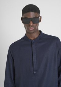Camicia a maniche lunghe blu navy con colletto alla band e dettagli con bottoni. Il modello indossa occhiali da sole neri oversize. Tessuto con una texture liscia.