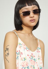 Óculos de sol retangulares pretos com acabamentos em metal, usados por uma pessoa com um vestido sem mangas de padrão floral e uma tatuagem no braço superior.