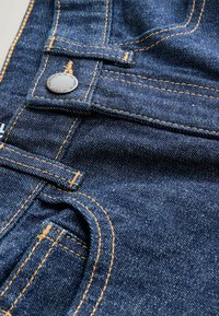 Jean en denim bleu foncé avec des coutures orange, un bouton en métal et un tissu texturé, mettant en avant les détails de la ceinture et des poches.