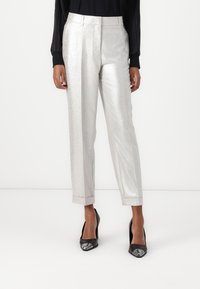 Pantalons argentés métalliques, taille haute, avec un devant plissé et des jambes fuselées ; associés à des talons noirs et un haut noir à manches longues plissé.