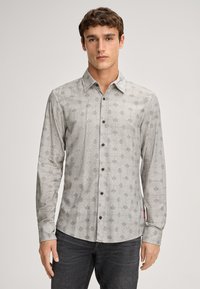 Jonge man in een lichtgrijs overhemd met lange mouwen en geometrische patronen, met donkere jeans, staande tegen een effen achtergrond.