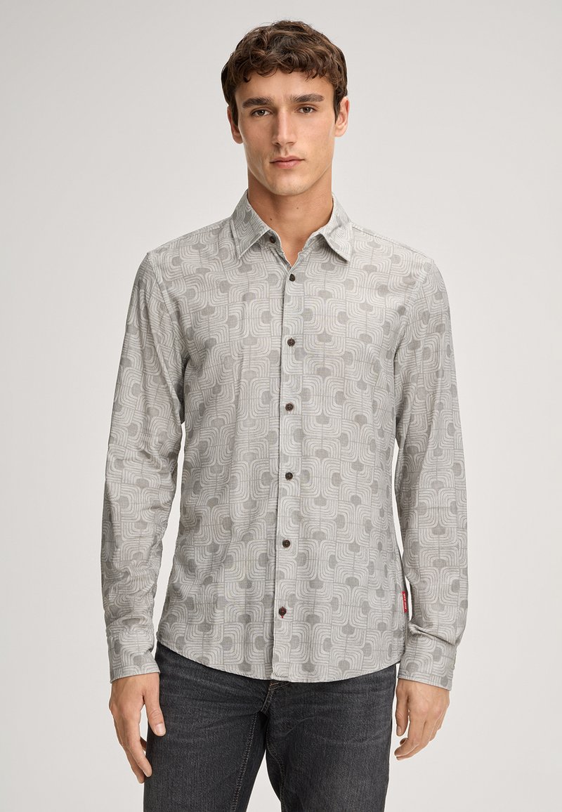 Jonge man in een lichtgrijs overhemd met lange mouwen en geometrische patronen, met donkere jeans, staande tegen een effen achtergrond.