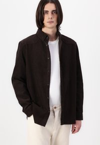 Camicia di velluto a coste marrone con design a bottoni, dotata di un colletto, tessuto testurizzato e bottoni neri, indossata sopra una camicia bianca.