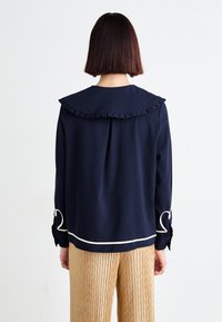 Ναυτική μπλούζα με ριγωτό κολάρο και λευκή τσιμπούκι; μακριά μανίκια με διακοσμητικές ζώνες. Φορεμένη με ανοιχτόχρωμες, υφασμάτινες, πλισέ παντελόνες.