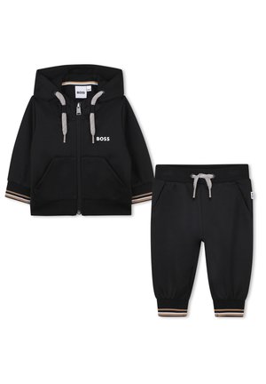 Veste noire à capuche avec fermeture éclair, poches avant et cordon de serrage. Pantalon assorti avec taille élastique et manchettes rayées. Les deux sont en matériau doux.