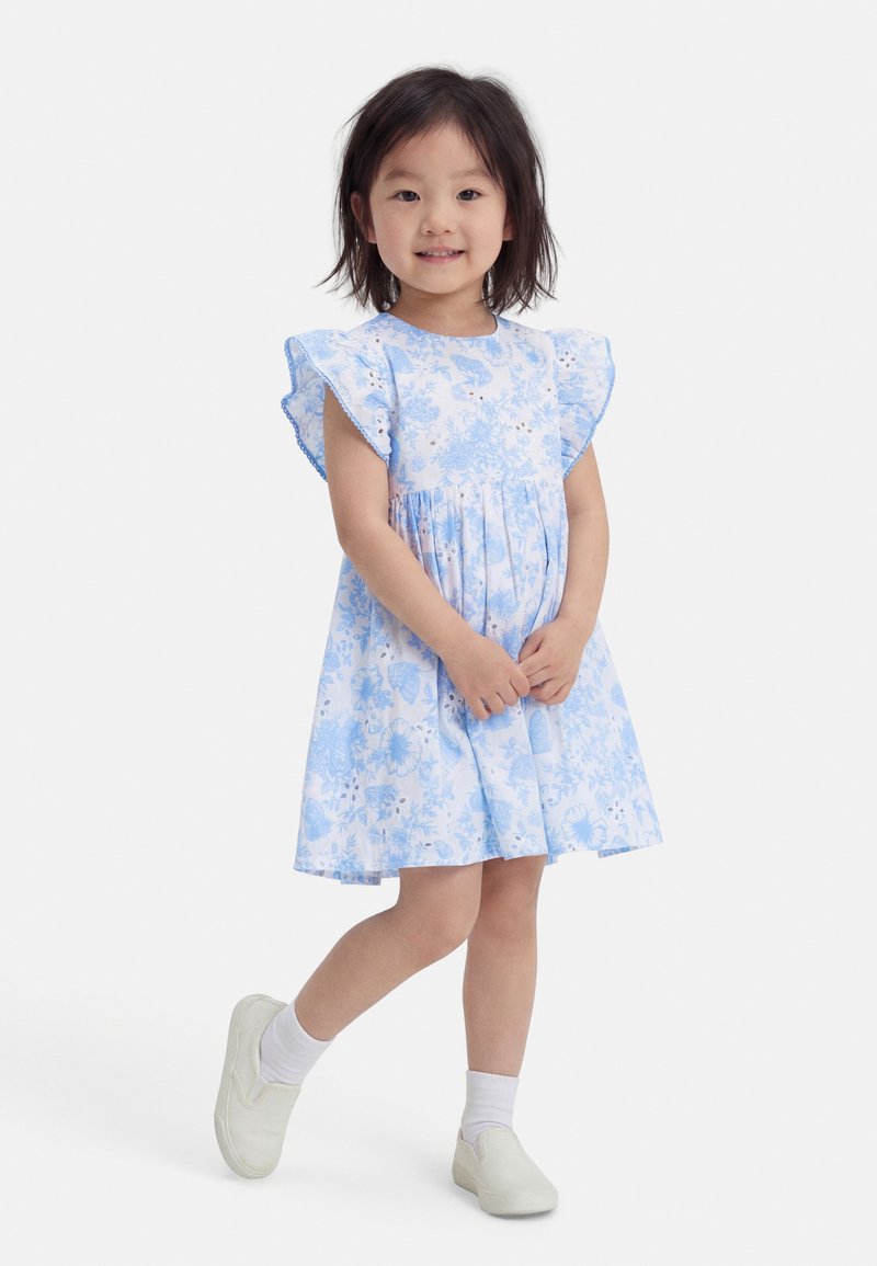 Νεαρή κοπέλα με μπλε και λευκό φόρεμα με λουλούδια και βολάν στα μανίκια, λευκές κάλτσες και παπούτσια slip-on, όρθια και χαμογελαστή.