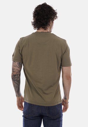 Hombre con cabello rizado que lleva una camiseta de manga corta verde oliva y jeans azules, mostrando la vista trasera con tatuajes en el brazo visibles.