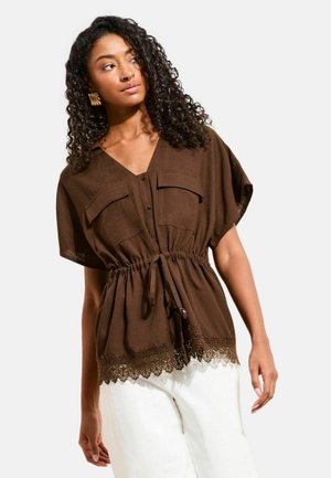 Žena s kučeravými vlasmi, oblečená v hnedom krátkoročnom topu na gombíky s opaskom so šnúrkou a čipkovaným lemom, doplneným bielymi nohavicami.