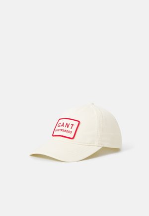 Witte baseballpet met een rood omrande patch met de tekst "GANT SHIRTMAKERS" op het voorpaneel tegen een effen achtergrond.