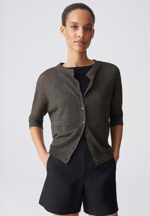 Frau mit kurzen Haaren, die eine dunkelolivfarbene, geknöpfte Strickjacke über einem schwarzen Top und schwarzen Shorts trägt und mit den Händen in den Taschen steht.