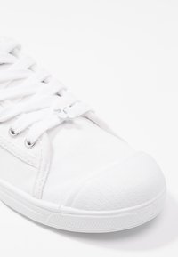 Basket en toile blanche avec une surface texturée, bout rond et lacets blancs. Présente des œillets en métal argenté et un détail de logo sur les lacets.
