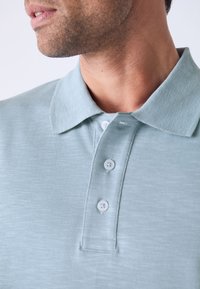 Polo vert clair en tissu texturé, doté d'un col et de quatre boutons blancs sur le devant, présentant un design décontracté.