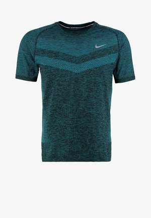 Jeune homme debout, bras croisés, portant un maillot de sport sombre à motifs, un short bleu à motifs et des chaussures de course noires.