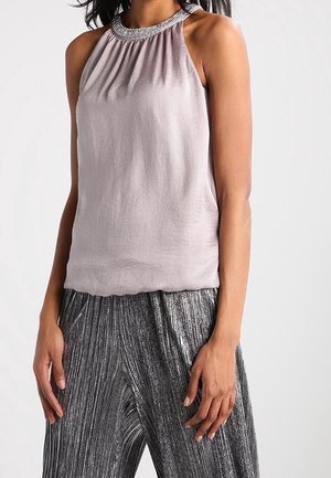 Femme portant un haut sans manches rose clair avec un col orné de perles argentées et un pantalon plissé métallique argenté taille haute, debout devant un fond blanc.