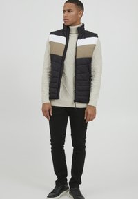 Gequiltete marineblaue Weste mit beigen und weißen horizontalen Streifen, getragen über einem hellen, strukturierten Pullover und schwarzen Hosen, kombiniert mit schwarzen Sneakers.