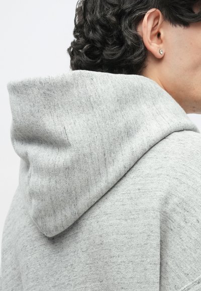 Persona con capelli ricci scuri che indossa una felpa con cappuccio grigio chiaro e un piccolo orecchino a perno nell'orecchio sinistro, vista di schiena rispetto alla fotocamera.