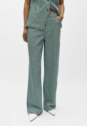 Femme portant un pantalon large à rayures verticales vertes et blanches avec un haut sans manches boutonné assorti, des chaussures blanches à bouts pointus et un bracelet argenté.