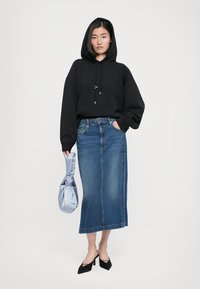 Ženska nosi črno kapuco, modri denim midi krilo, črne pete in drži svetlo modro satensko torbico na enobarvni beli ozadju.