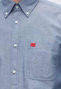 Ανοιχτό μπλε πουκάμισο με κουμπιά και μπροστινή τσέπη με μικρή κόκκινη ετικέτα που αναγράφει «HUGO».