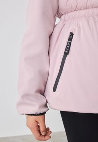 Lyserød jakke med elastiske manchetter, indsnævret talje og sidelomme med lynlås. Glat, mat tekstur; har branding på lynlåshanken.