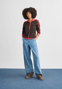 Black zip-up jacket with red accents and three stripes; paired with light blue wide-leg jeans and brown sneakers.