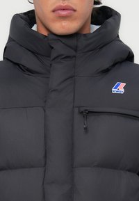 Doudoune noire avec un col haut, dotée d'une poche zippée et du logo distinctif K-Way. Le tissu a une finition lisse et mate.