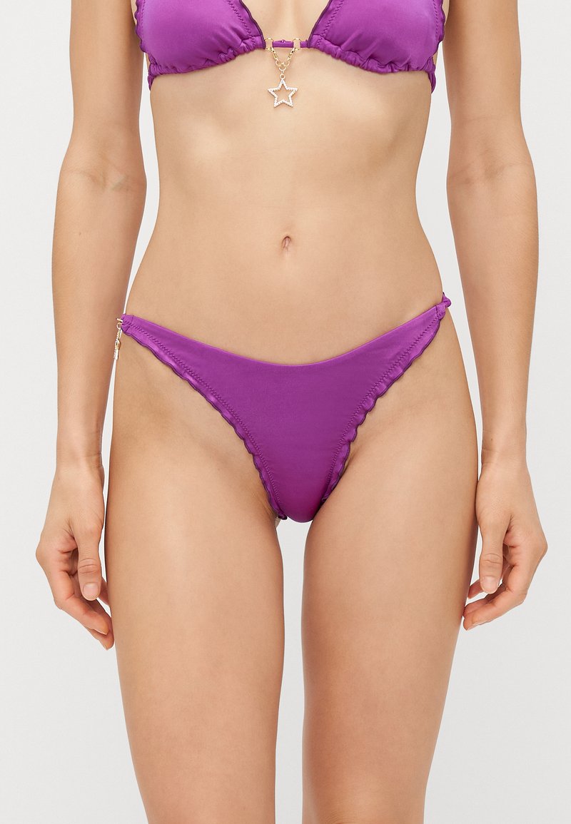 Mulher vestindo um biquíni roxo, com parte de baixo e parte de cima combinando, com um pendente em forma de estrela entre os copas, de pé contra um fundo simples.