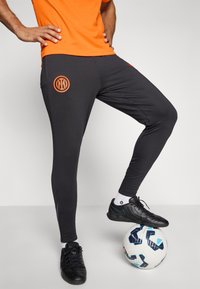 Melnās sporta bikses ar oranžu logo, pieguļošs dizains un sānu akcenti. Apvienotas ar melnām futbola kurpēm uz rakstainas baltas bumbas.