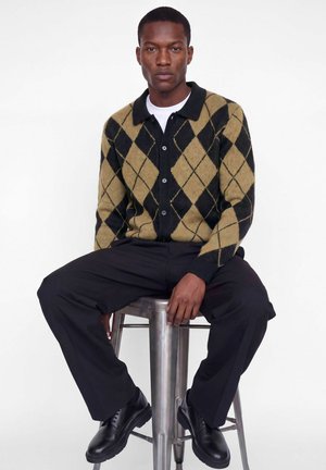 Homme assis sur un tabouret en métal portant un cardigan argyle marron et noir, un pantalon noir, des chaussures noires et une chemise blanche sur un fond uni.