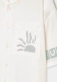 Camisa blanca de manga corta con botones y bolsillo frontal que presenta un diseño abstracto de sol gris y un estampado geométrico parcial en las mangas.