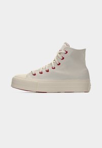 Μπεζ πάνινο αθλητικό παπούτσι τύπου high-top με χοντρή σόλα, κόκκινα μεταλλικά οπές για κορδόνια και κορδόνια σε ίδιο χρώμα, σε ανοιχτό γκρι φόντο.