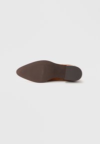 Chaussure à talon en daim marron avec un bout pointu et une semelle en caoutchouc texturée, présentant une bordure lisse et une marque subtile au bas.