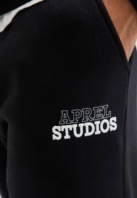 Sorte svedbukser lavet af bomuld med en broderet hvid tekstlogo "APREL STUDIOS" på venstre lår. Har en afslappet pasform og sidelommer.