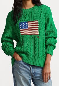 Personne portant un pull vert en maille torsadée avec un motif tricoté du drapeau américain, associé à un jean bleu clair, main dans la poche.