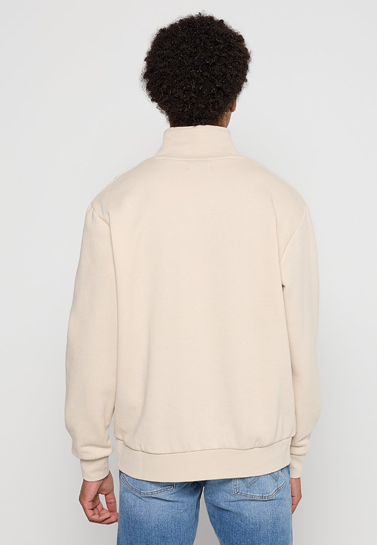 Sudadera beige con cuello alto y puños de canalé. La tela parece suave y texturada. Presenta un corte relajado y detalles de costura sutiles.