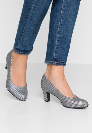 Pieds chaussés de escarpins à talons bas à motif pied-de-poule noir et blanc, associés à un jean bleu raccourci, debout sur un sol gris.