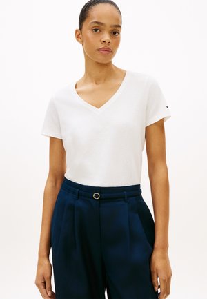 Vrouw met haar in een knot, draagt een wit T-shirt met V-hals en korte mouwen en marineblauwe, hooggetailleerde broek met een smalle riem, staand tegen een effen achtergrond.