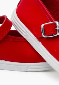 Zapatos sintéticos rojos con suela de goma blanca, que presentan una correa y un detalle de hebilla plateada. Textura suave con forma redondeada.